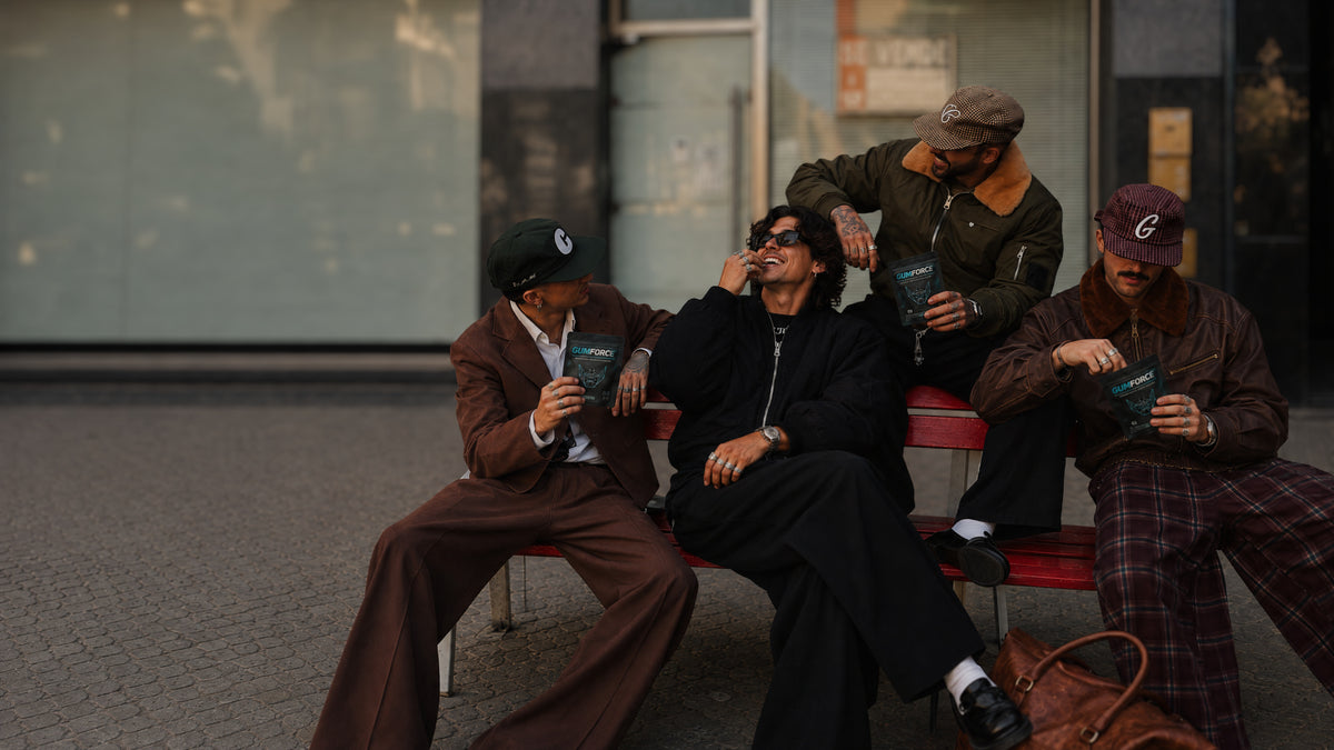 Quatre hommes sur un banc, sachets de chewing-gums GUMFORCE en main, l'un met un chewing-gum en bouche
