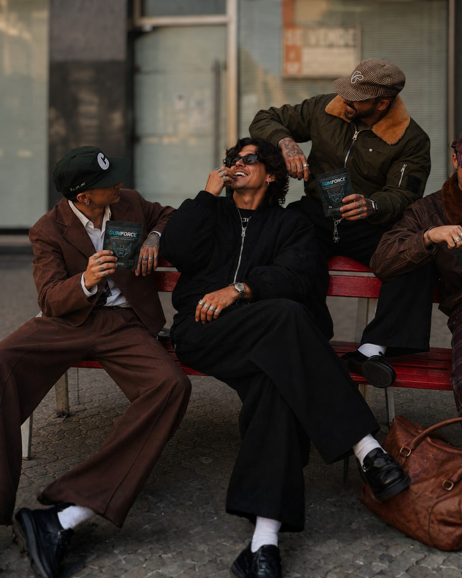 Quatre hommes sur un banc, sachets de chewing-gums GUMFORCE en main, l'un met un chewing-gum en bouche